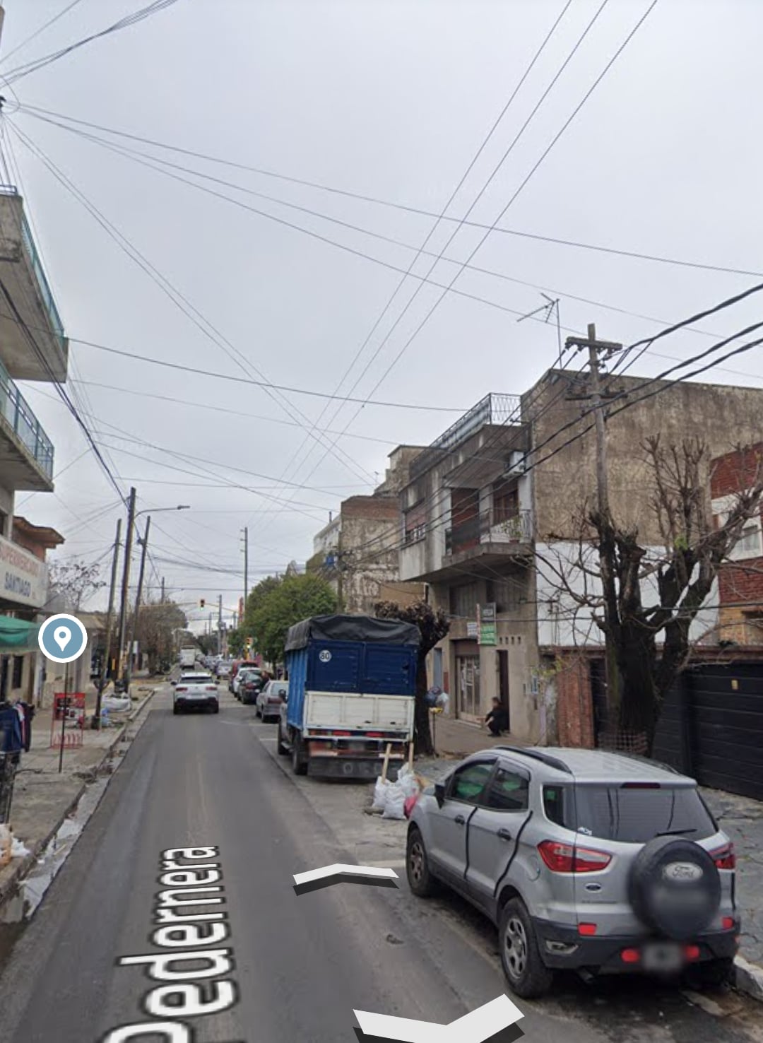 A quiet street scene is depicted, featuring vehicles parked along both sides. A blue truck is noticeable near the center, while a variety of buildings line the street. Utility poles and overhead wires are visible against a gray sky.