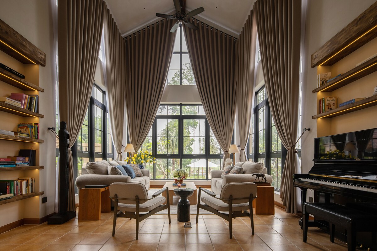 A spacious living area features high ceilings with large windows, allowing natural light to fill the space. Soft-colored sofas are complemented by wooden side tables, while shelves lined with books provide a cozy touch. A piano stands in the corner, enhancing the room's inviting atmosphere.