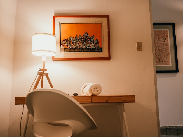 A bright workspace is presented with a wooden desk and a white chair. A lamp stands on the desk beside neatly rolled towels. An orange artwork highlights the wall, while an adjacent doorway leads to another room, creating a sense of depth in the space.