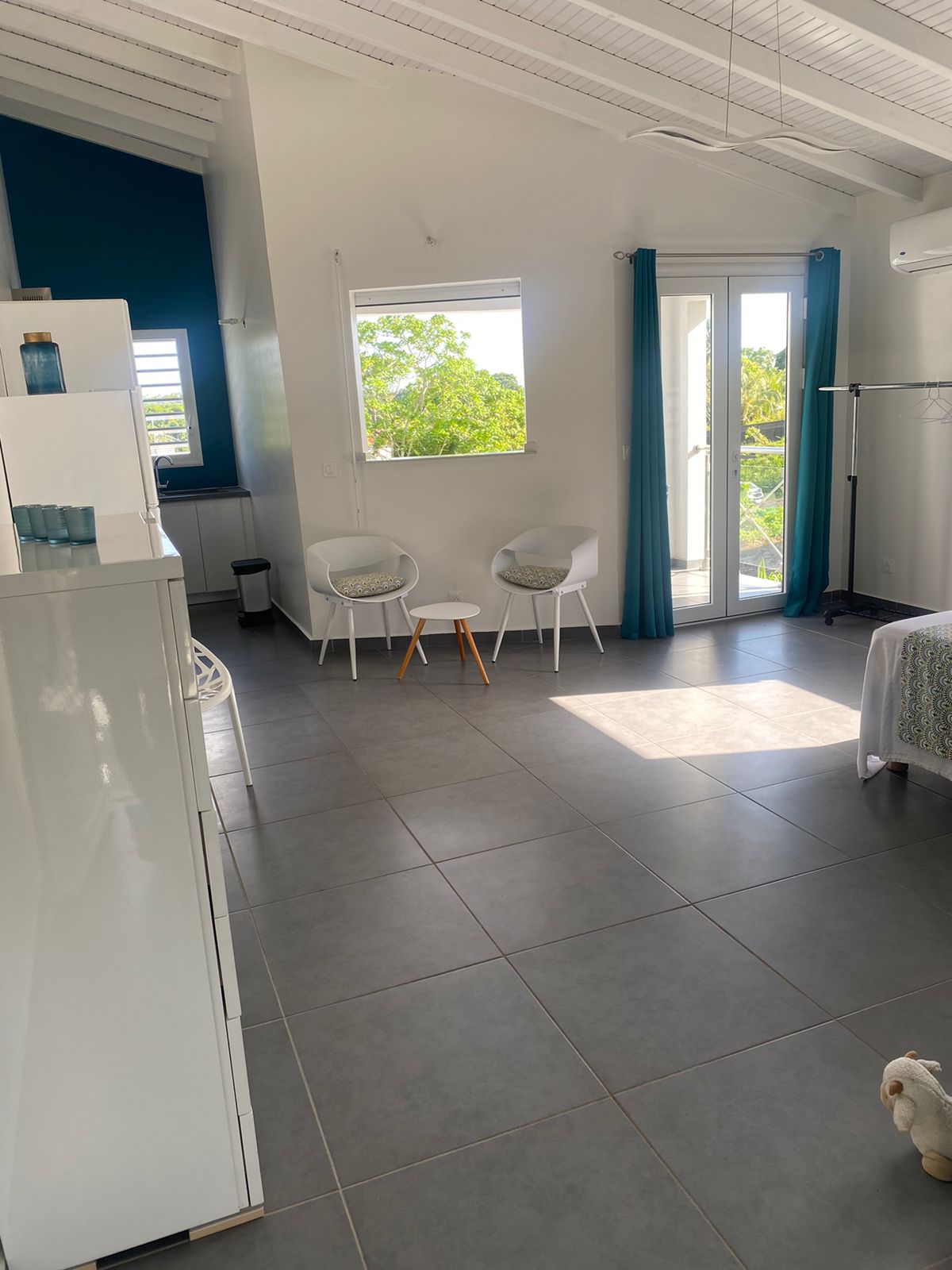 A spacious interior is showcased with sleek tiled flooring and high ceilings. Two modern chairs are positioned beside a small table, with a doorway leading to a balcony. Natural light filters through the curtains, highlighting the clean design and vibrant accents.