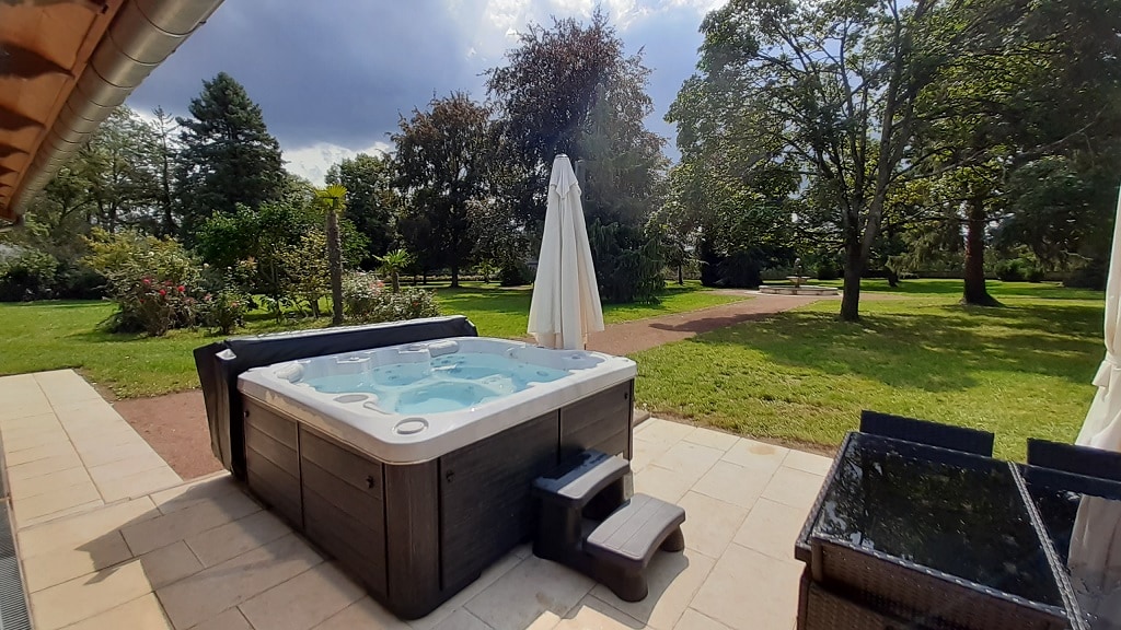 A heated spa is positioned on a tiled terrace, surrounded by a well-maintained park. Lush greenery is visible in the background, with trees and flower beds enhancing the natural setting. A shade umbrella stands nearby, adding comfort to the outdoor space.