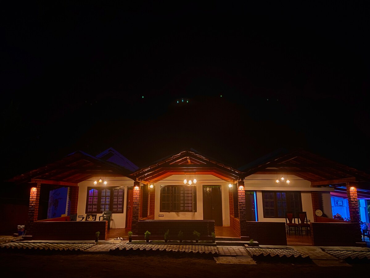 The exterior of a bungalow is illuminated at night, highlighting the intricate wooden architecture. Ambient lighting enhances the welcoming atmosphere, while the surrounding landscape remains softly shadowed by the night sky. A spacious porch features seating arrangements, providing an inviting entry point.