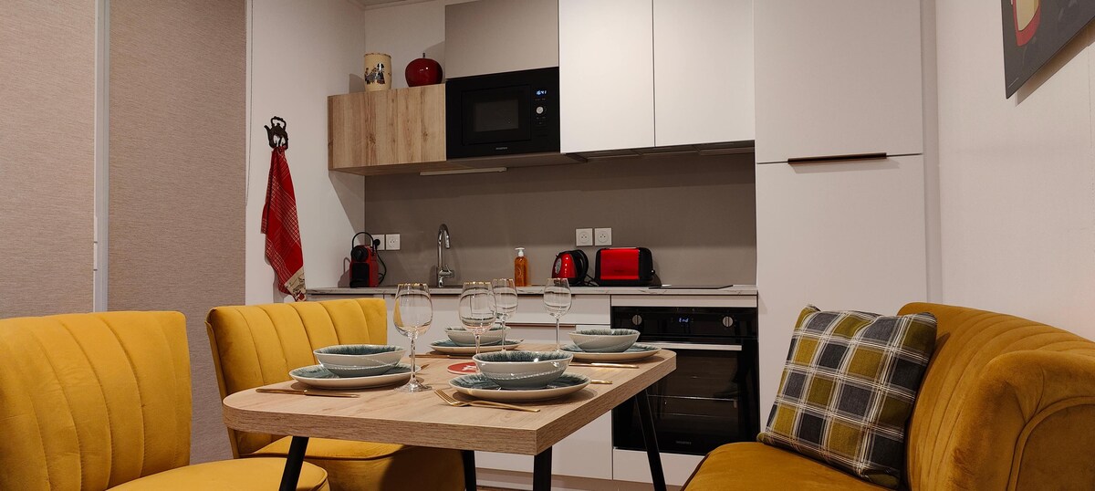 A modern kitchen area is presented with a wooden dining table set for four, featuring plates and glasses. Yellow sofas flank the table, complementing the cabinetry and appliances, including a microwave and toaster. A cozy yet functional atmosphere is conveyed by the arrangement.