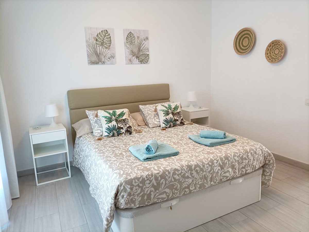 A well-lit bedroom features a spacious bed adorned with light-colored linens and decorative pillows. Two framed botanical prints are displayed above the headboard, complemented by woven wall decor. Nightstands with lamps are positioned on either side of the bed.