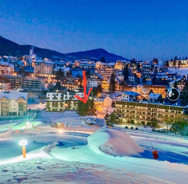 A snowy landscape is captured at dusk, with soft blue tones enveloping the scene. The charming architecture of nearby buildings is visible, including a residential structure partially obscured by snow. Illuminated paths and sloped terrain demonstrate the winter setting, enhancing accessibility for outdoor activities.