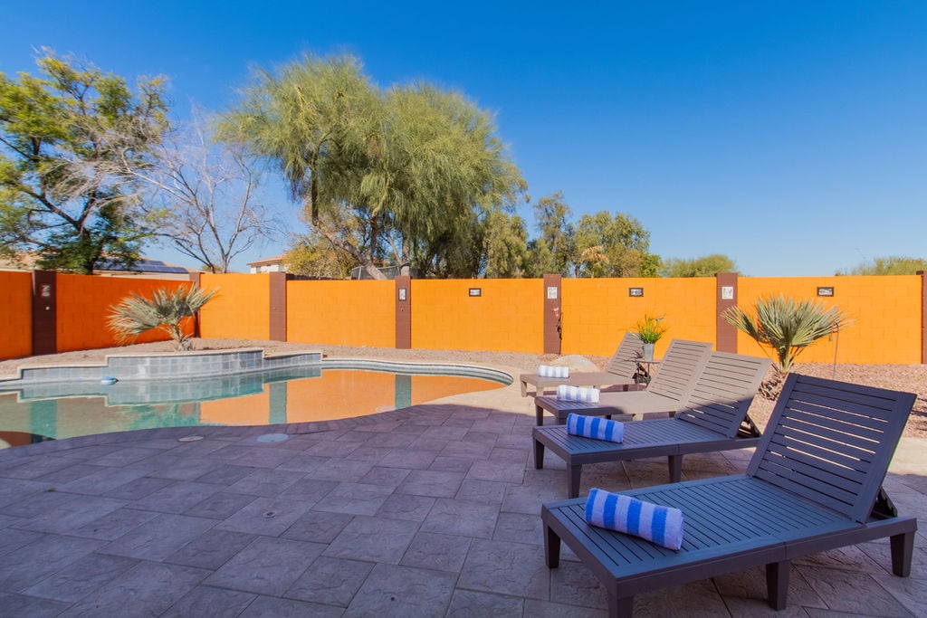 An inviting backyard features a spacious swimming pool, bordered by a sun terrace with multiple lounge chairs. Soft towels are neatly arranged, and vibrant orange walls add a pop of color against the clear blue sky.