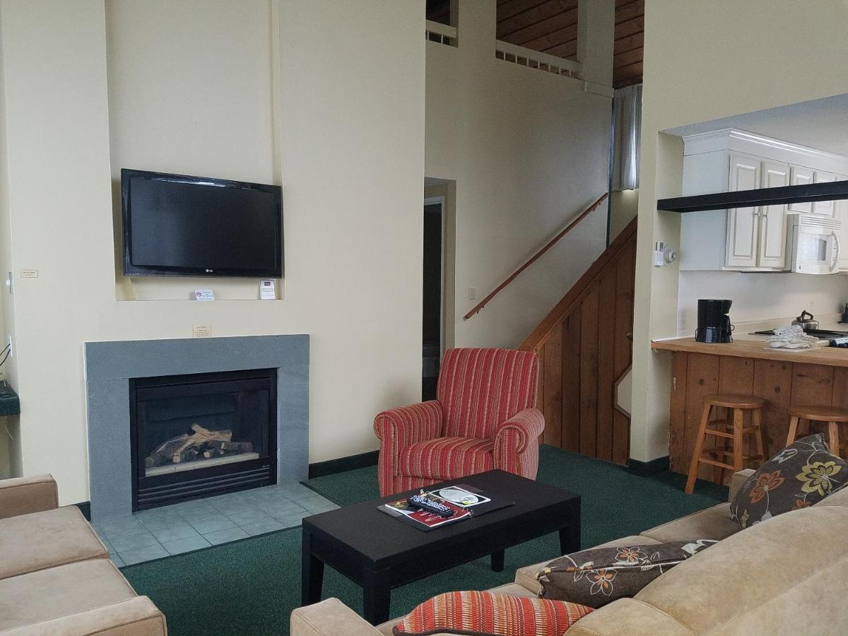 A living area is presented with a cozy seating arrangement, featuring a patterned armchair and a neutral-colored sectional sofa. A modern fireplace is situated beneath a wall-mounted television. The kitchen area and dining space are visible in the background, enhancing the openness of the setting.