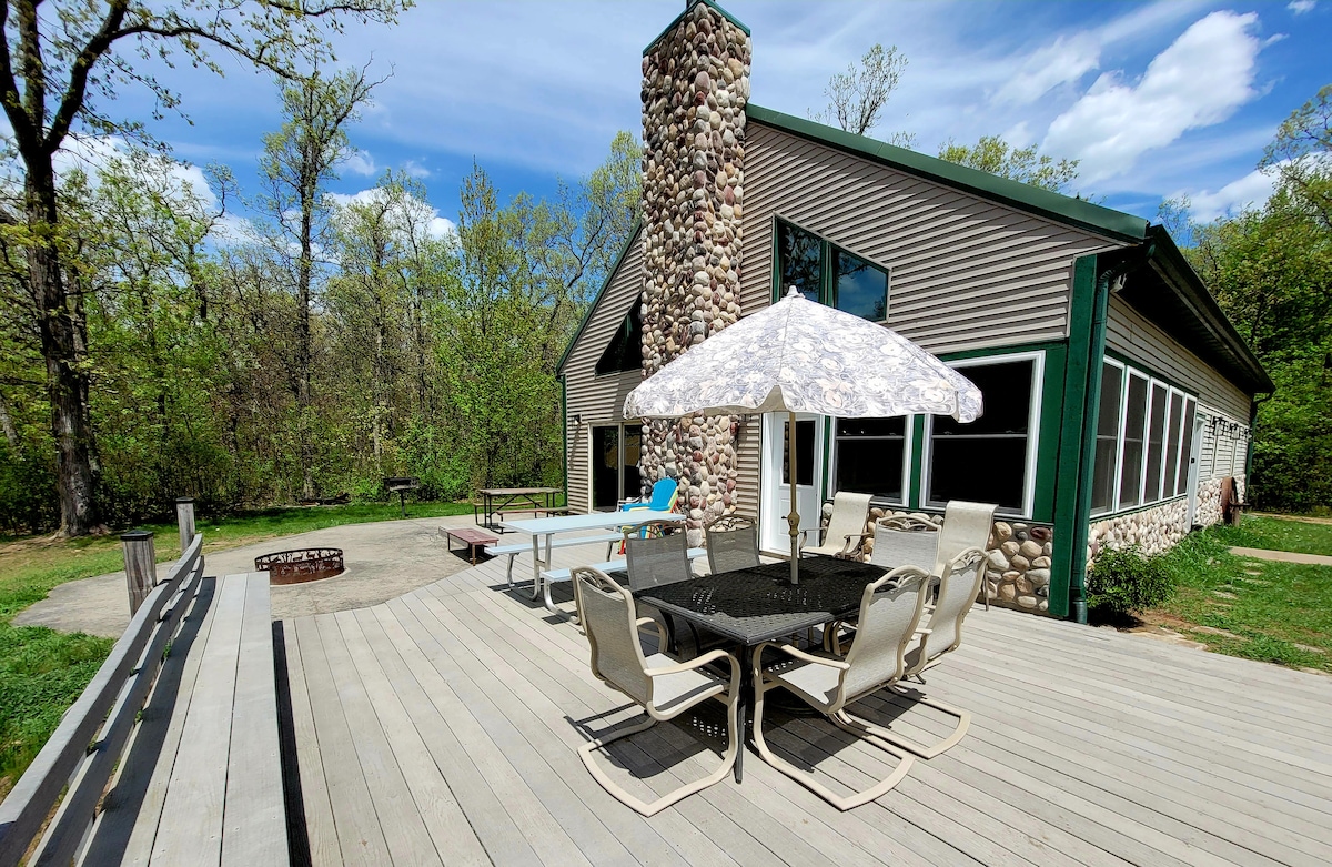 A spacious outdoor deck includes a large table with an umbrella, surrounded by multiple chairs. A fire pit is visible nearby, along with a ping pong table. The cabin's exterior features a stone chimney, and green accents complement the natural wooded surroundings.