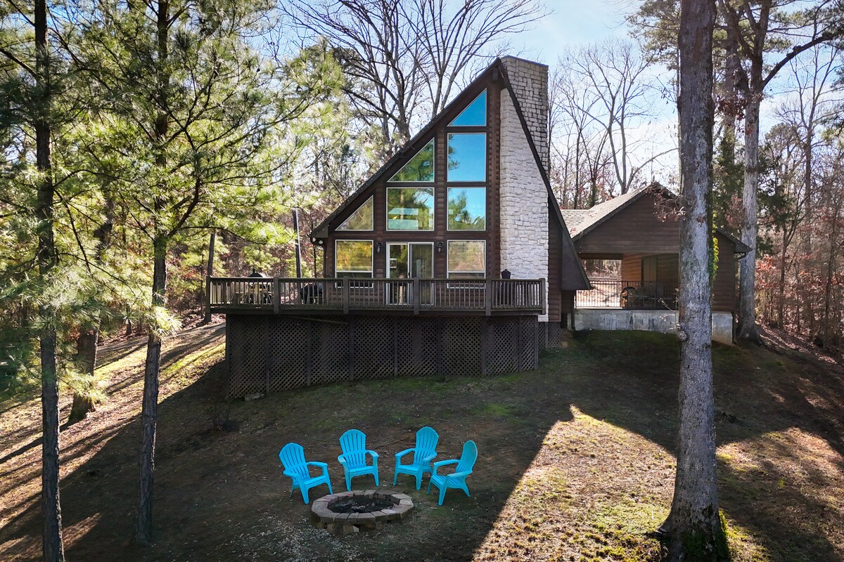The A-frame cabin is surrounded by tall pines, showcasing large glass windows that reflect natural light. A wrap-around deck offers seating with blue Adirondack chairs positioned around a fire pit, inviting relaxation in a serene outdoor setting.