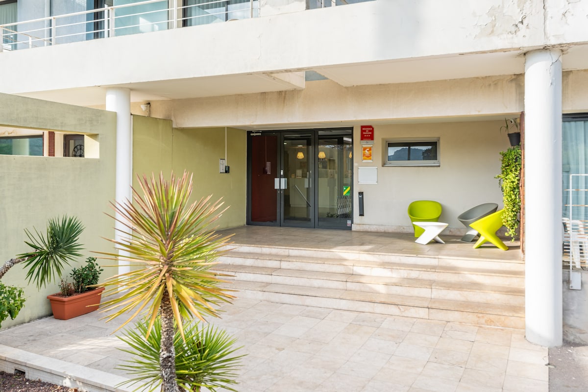 The entrance to the building features a set of stair steps leading to a glass door surrounded by greenery. Two modern chairs in vibrant colors are positioned on the porch, inviting relaxation in the open space.