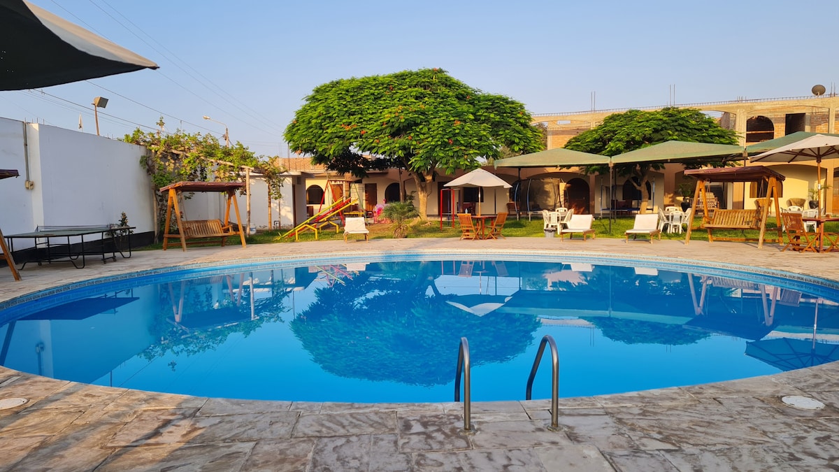 A refreshing swimming pool is surrounded by well-maintained gardens, with lounge chairs and shaded seating areas nearby. The clear blue water reflects the vibrant green foliage of a large tree, providing a serene setting for relaxation and family activities.