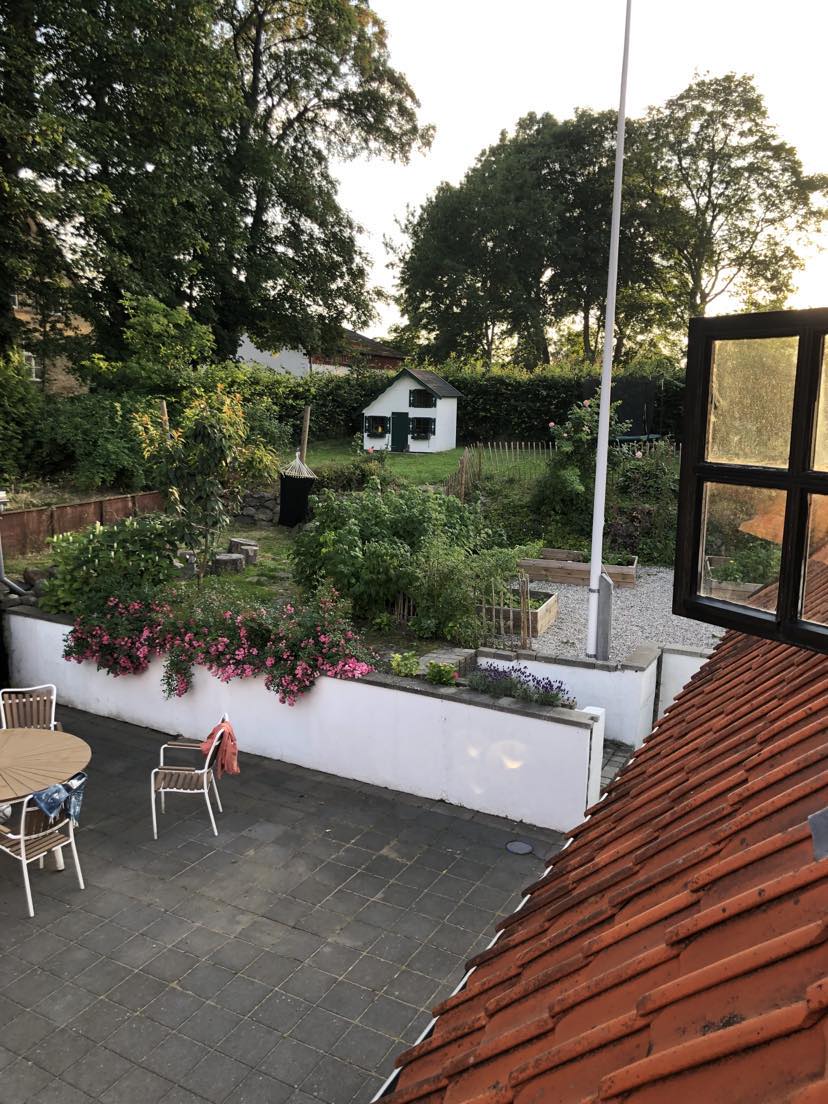 A garden area is visible from an upper window, featuring green plants and colorful flowers along a white wall. A small shed stands in the background, while a round table with chairs is situated on a stone patio, inviting outdoor gatherings.