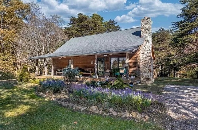 The charming cabin is set amidst lush greenery, featuring a covered porch adorned with seating. A stone chimney rises from the side, complementing the rustic wooden facade. Vibrant flowers bloom in front, enhancing the natural beauty of the serene surroundings.