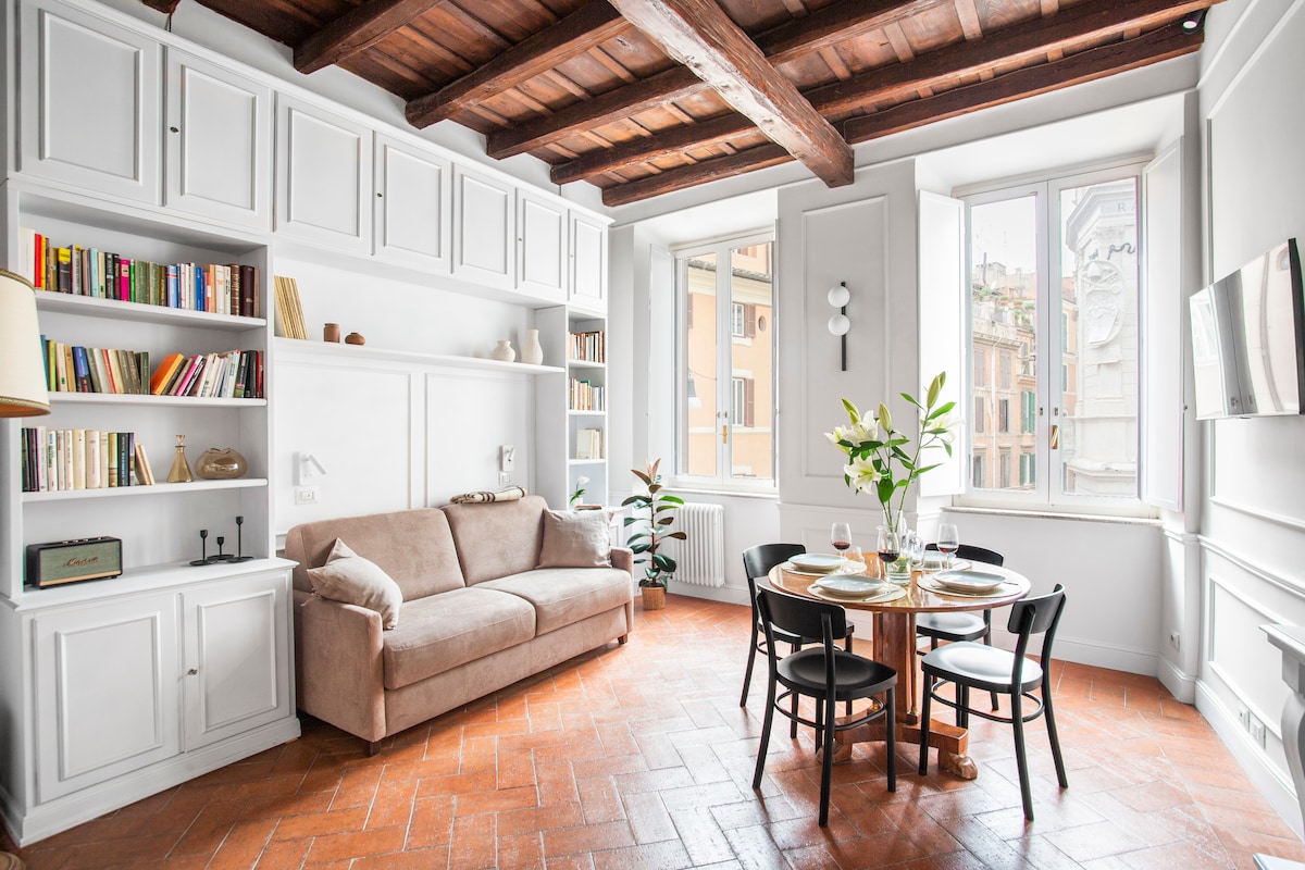 A well-lit living area features original wood beams and terracotta flooring. A sofa is positioned beside a circular dining table set for two, with elegant tableware. Floor-to-ceiling windows allow natural light to illuminate the space, offering views of the vibrant street outside.