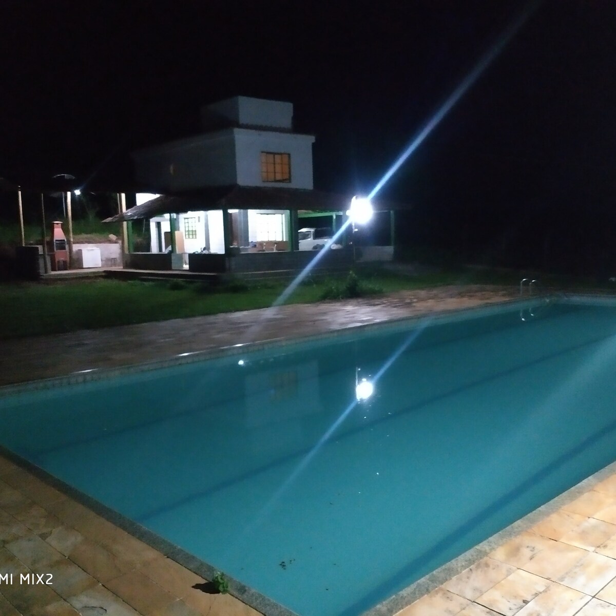 A serene pool area is illuminated by soft lights against the night sky, reflecting on the water's surface. A spacious house can be seen in the background, with large windows highlighting the inviting ambiance of the outdoor space.