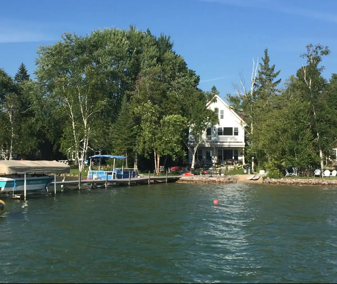 A lakefront property is visible with a three-story house surrounded by lush green trees. A sandy beach is accessible from the backyard, and a dock with a boat and seating area is seen nearby. The tranquil water reflects the clear blue sky.