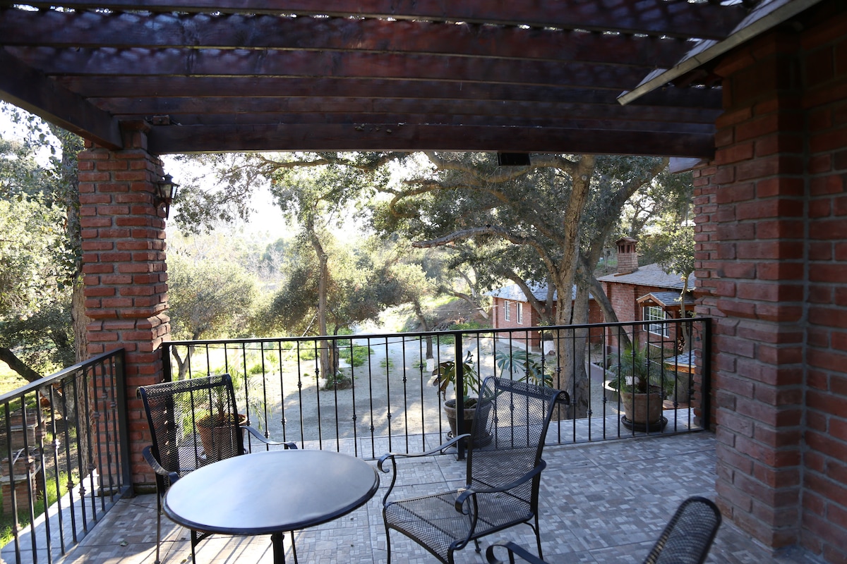 A shaded patio area is seen, featuring a round table and four metal chairs. The space is surrounded by greenery and overlooks a pathway leading to separate units in the distance. The wooden pergola provides shelter, creating a serene outdoor setting.