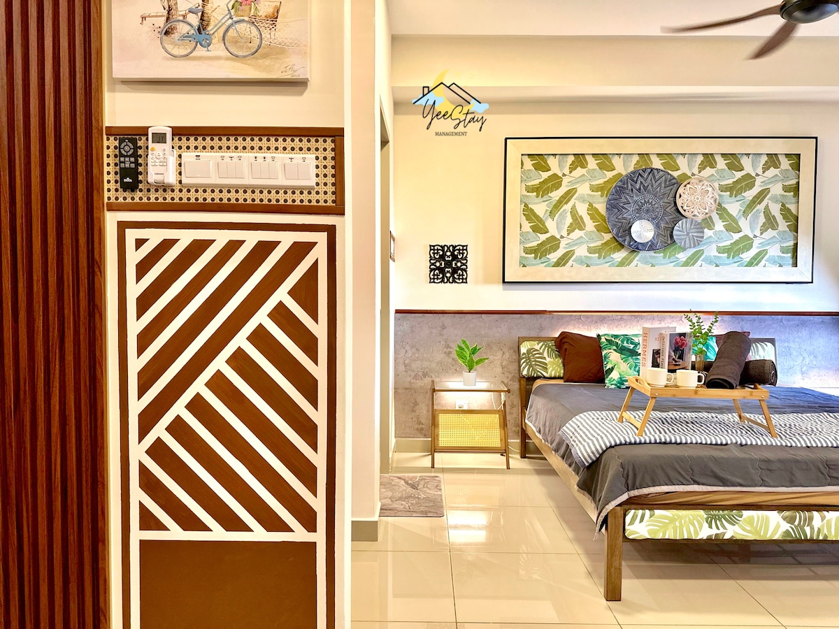 A spacious room features a comfortable bed adorned with decorative pillows and a tray positioned atop it. The walls display botanical artwork, and natural light is welcomed through a nearby window. A stylish accent wall provides texture, enhancing the overall design.