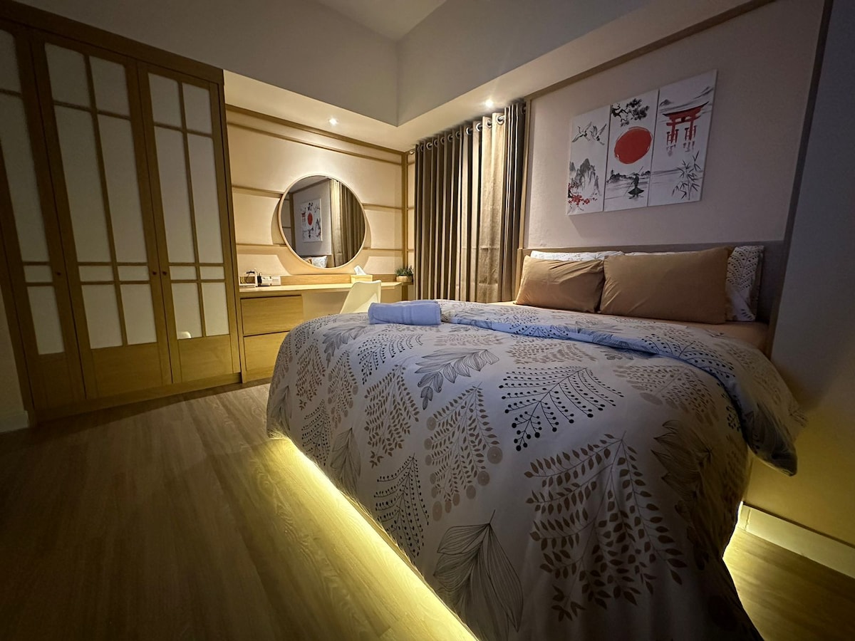A spacious bedroom features a large bed with soft linens and an illuminated base. A large mirror and a wooden desk are positioned in front of the bed, while a wardrobe with sliding doors is visible in the background. Soft lighting enhances the serene atmosphere.