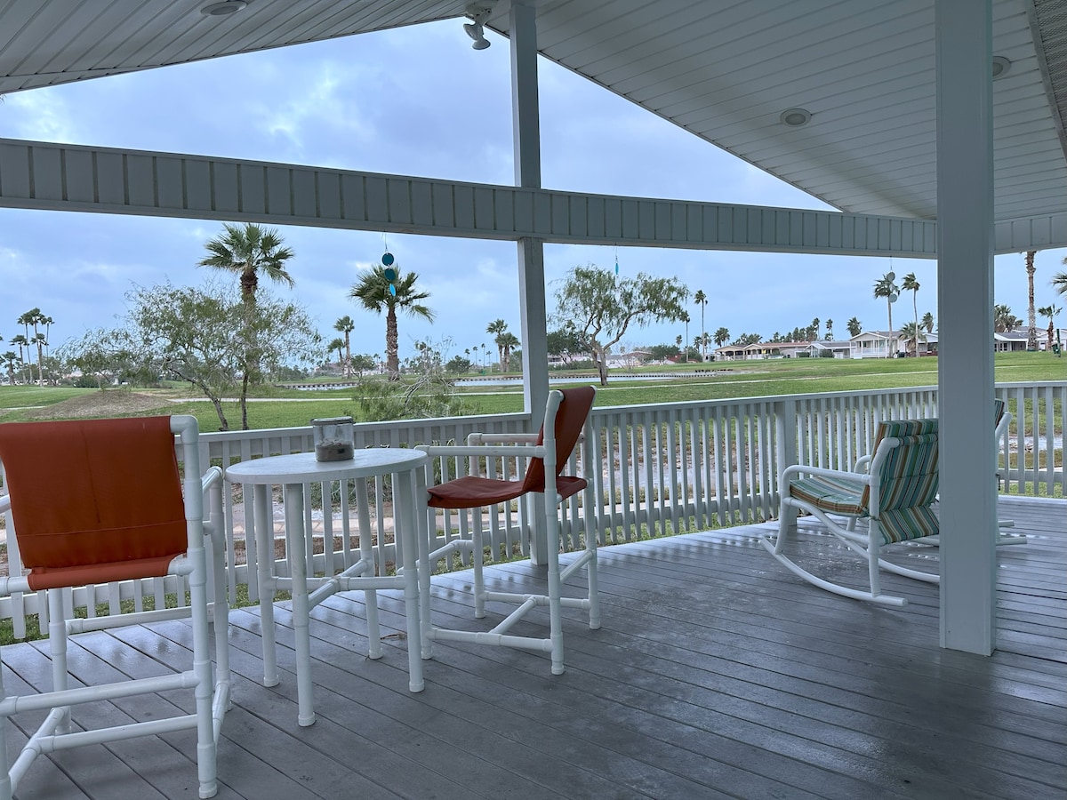 A covered patio features a table and four outdoor chairs, with views of palm trees and green spaces. The flooring is a light-colored wood, and overhead lighting is embedded in the ceiling. Surrounding railings provide a sense of openness to the outdoor environment.