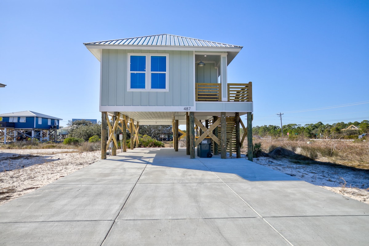 A newly built beach house is elevated on stilts, featuring light green siding and a metal roof. Two large windows are visible on the front, allowing ample natural light. A concrete driveway extends in front, providing easy access. Surrounding sandy areas are dotted with low coastal vegetation.