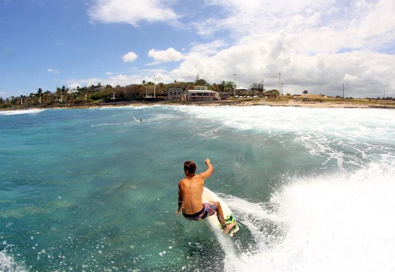 A surfer rides a wave in clear blue waters, with a shoreline visible in the background. The sky contains scattered clouds, and greenery is noticeable on the coastal land, suggesting a vibrant outdoor setting. The energy of the waves creates dynamic movement in the image.