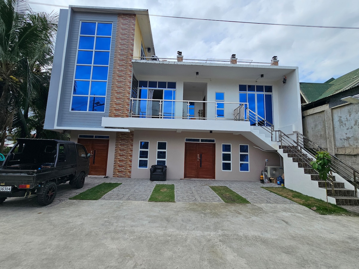 The exterior of the modern building features large blue-tinted windows that reflect the sky. A staircase leads to the upper level, and the entrance is framed by two wooden doors. Paved pathways and landscaped grass areas create an inviting approach.