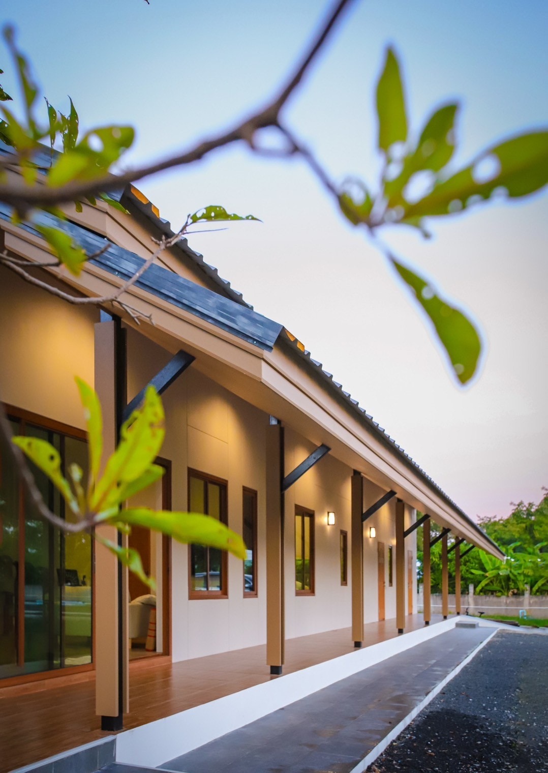 The exterior of a modern building is framed by lush green leaves. The structure features a sloped roof and multiple large windows, allowing natural light to illuminate the light-colored walls. A pathway runs alongside the building, leading to a landscaped area.