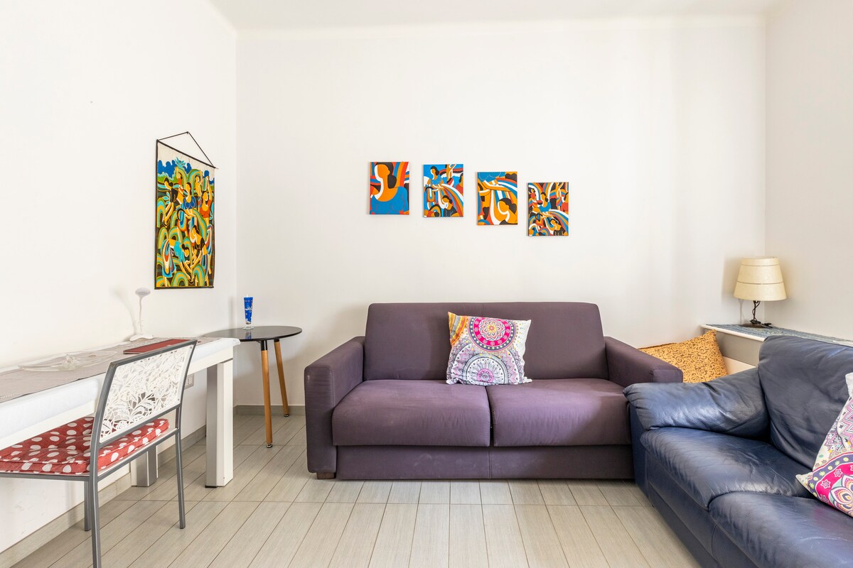 A cozy living area showcases a dark purple sofa and a navy blue leather couch. Colorful artwork is displayed on the white wall, while a small round table and a dining table with decorative chairs provide seating options. Natural light brightens the space.
