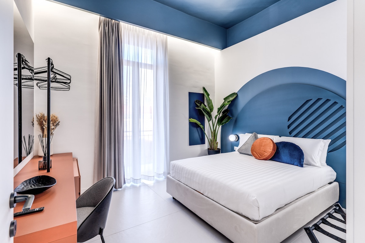 A modern bedroom features a spacious bed with a white striped duvet and decorative pillows. A vibrant blue accent wall adds visual interest. Natural light enters through sheer curtains, illuminating the room. A stylish desk and chair are positioned beside the bed, complemented by a potted plant.
