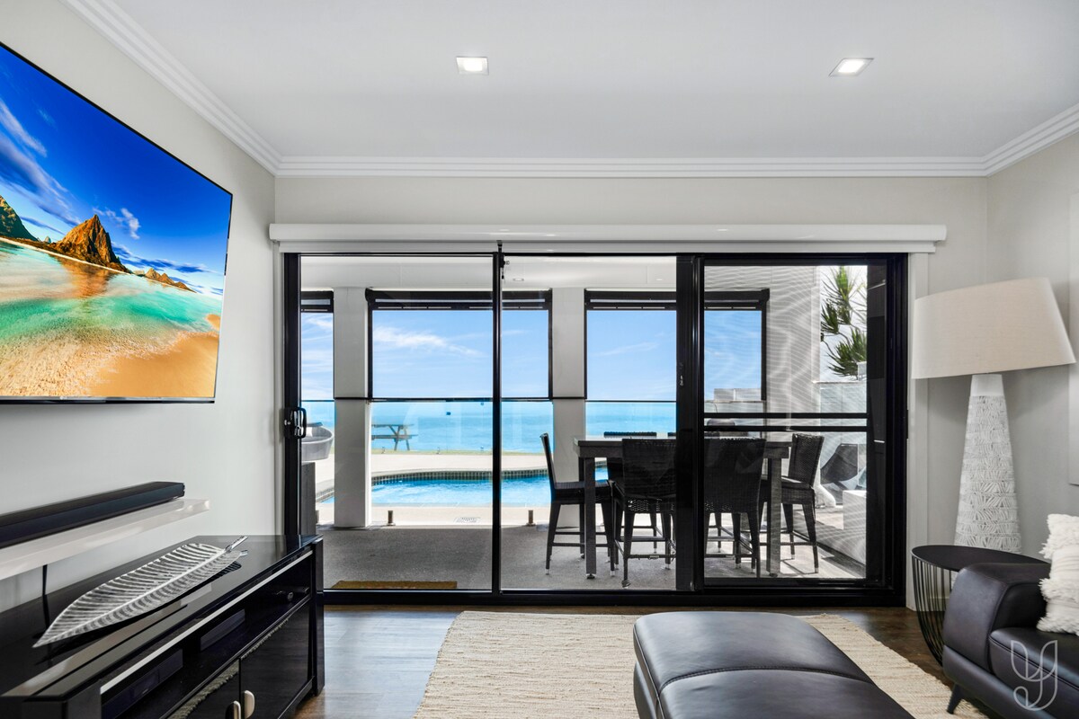 A spacious living area features large glass doors that open to a beautiful ocean view. A modern TV is mounted on the wall, and a comfortable seating arrangement is positioned beside a sleek lamp. Natural light enhances the inviting atmosphere.