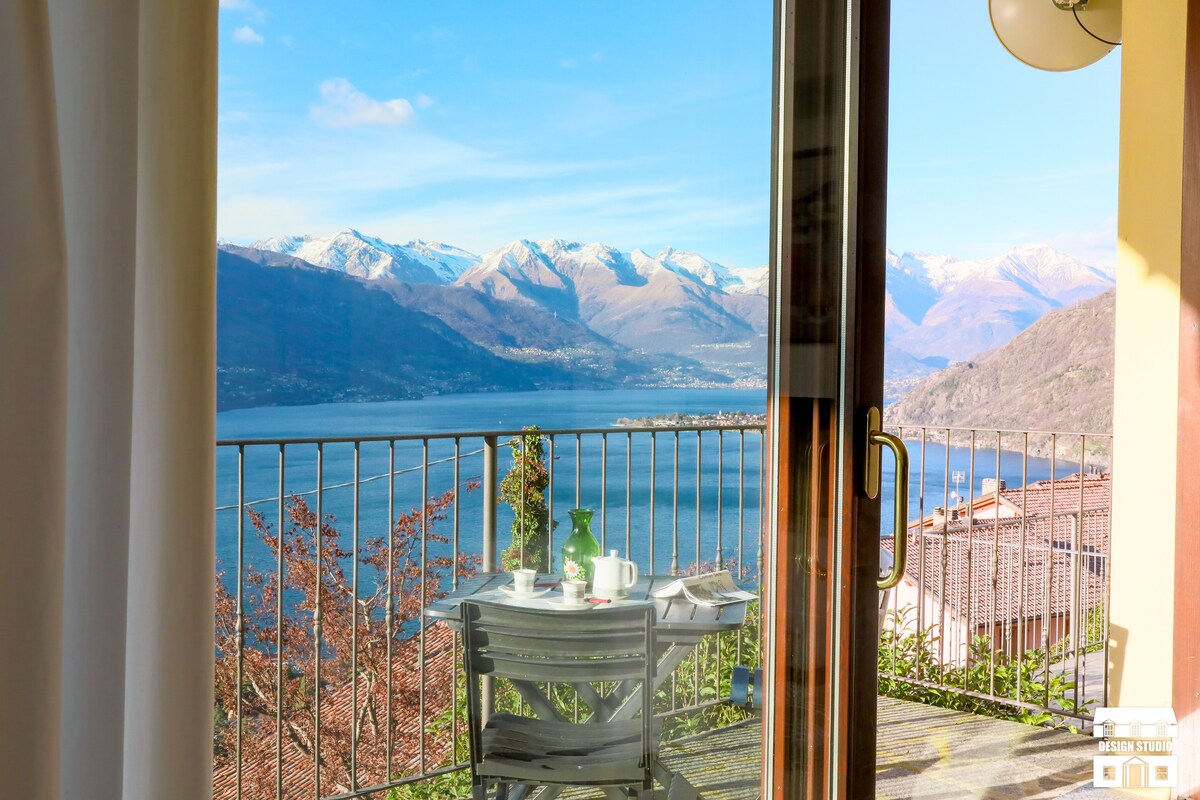 A scenic view of Lake Como is framed by a glass door, revealing a balcony with a small table and two chairs. The tranquil waters and surrounding mountains create a serene backdrop, while the blue sky and sunlight enhance the overall atmosphere.