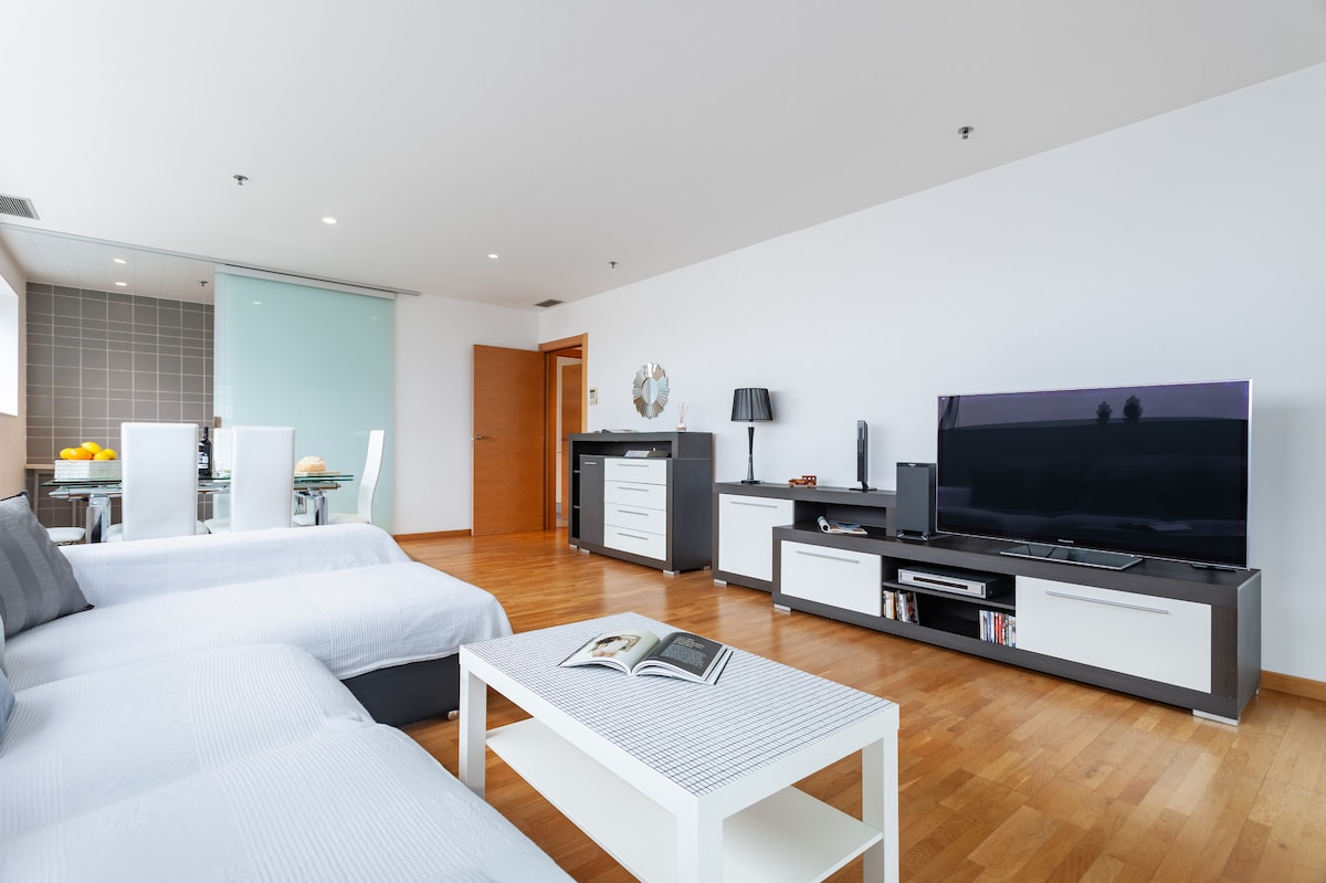 An open living area features a sleek couch and a contemporary coffee table with a magazine on top. A large television sits on a minimalist entertainment center. A dining space is visible in the background, complemented by glass sliding doors and wood flooring.