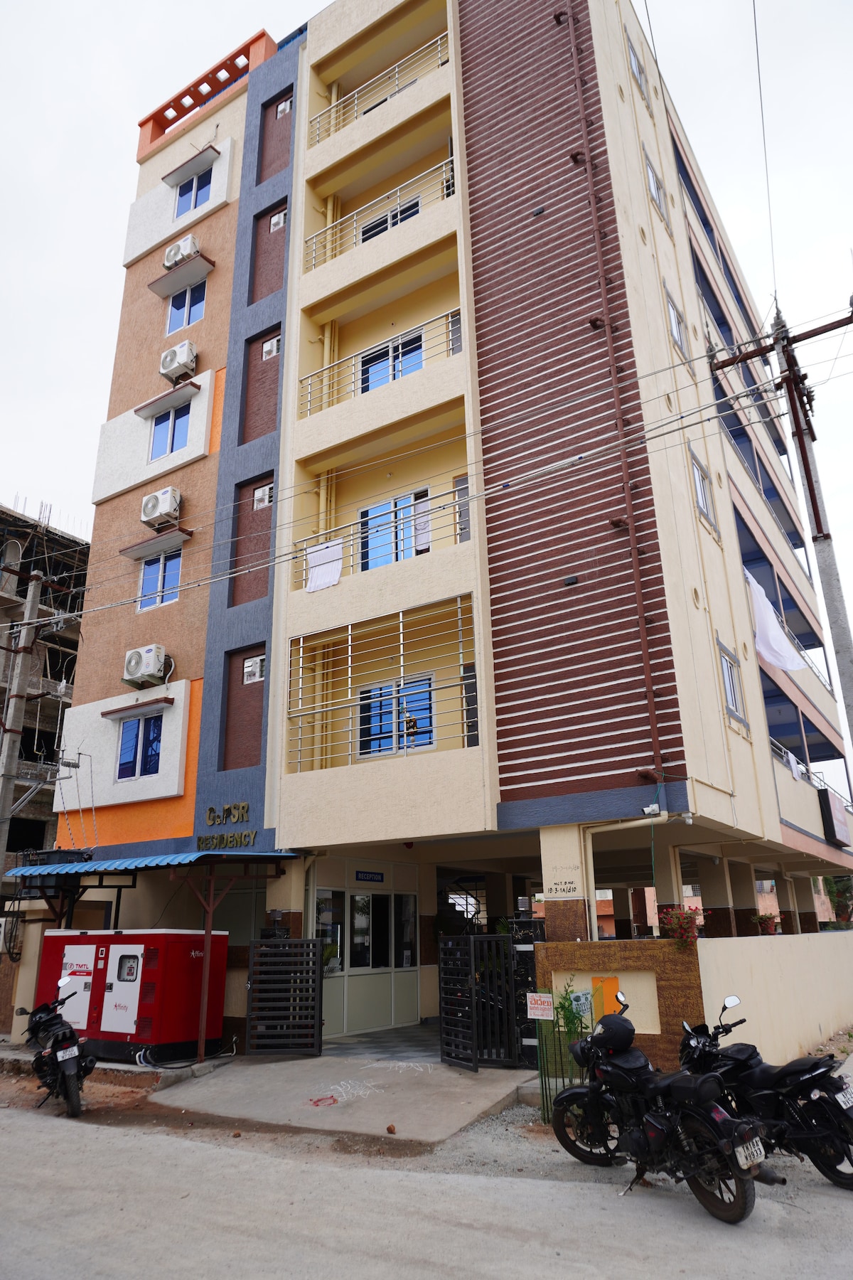 The exterior of a modern four-story building is captured, featuring a blend of earthy tones and blue accents. Multiple balconies are visible, with air conditioning units mounted on the walls. A secure entrance is accessed through a gated area, with parked motorcycles situated nearby.