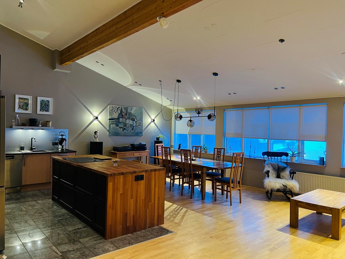 An open-concept kitchen and dining area features a large wooden island, modern appliances, and a spacious dining table surrounded by chairs. Natural light enters from multiple windows, highlighting the warm tones of the wooden accents and the sleek flooring.