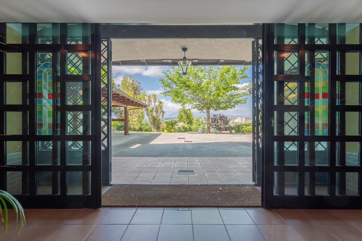 The entrance showcases large glass doors with decorative patterns, leading to an open area with natural light flooding in. A view of a lush garden and outdoor space is visible, framed by greenery and patio elements, creating a welcoming transition from indoors to outdoors.