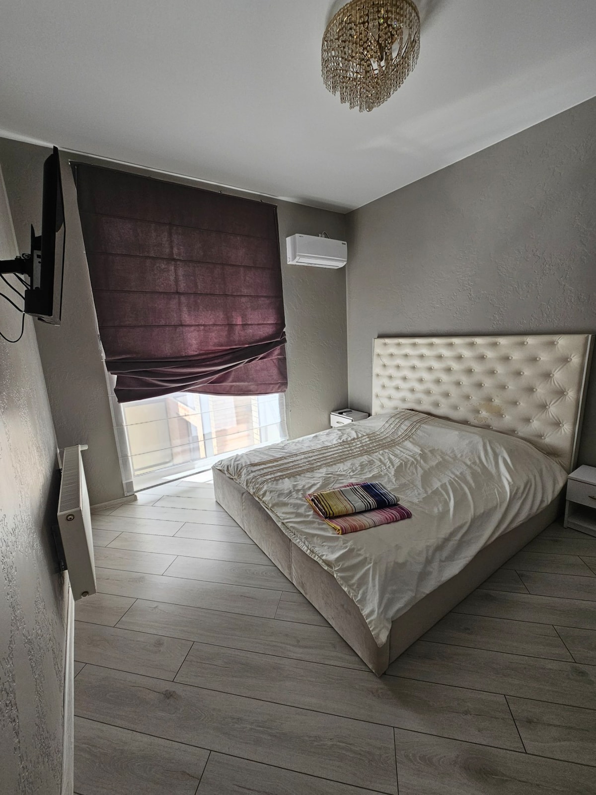 A well-appointed bedroom features a spacious bed with a tufted headboard. A wall-mounted television is present, and a large window is covered by dark, relaxing blinds. Light wood flooring complements the neutral wall tones and adds to the room's contemporary design.