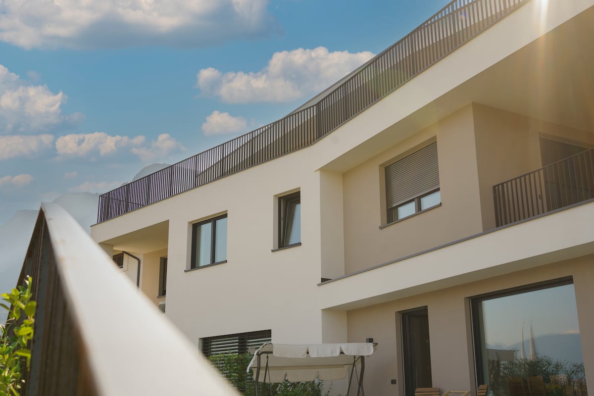 A modern apartment building features a light-colored facade with multiple windows and balconies. A railing extends in the foreground, leading to a terrace that offers a scenic view. Soft clouds are visible against a bright blue sky.