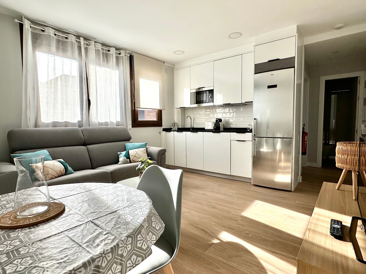 A bright and modern living area features a sleek gray sofa complemented by decorative cushions. A dining table is set with a patterned tablecloth. Contemporary kitchen appliances are visible in the background, along with large windows that allow natural light to fill the space.