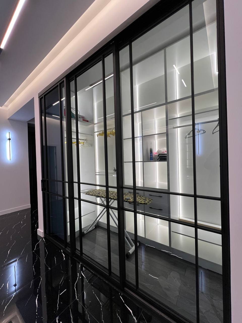 A modern closet features sleek glass doors with black framing. Inside, illuminated shelving displays organized storage, including a hanging space and an ironing board. The flooring reflects the design, enhancing the sleek and polished appearance of the space.