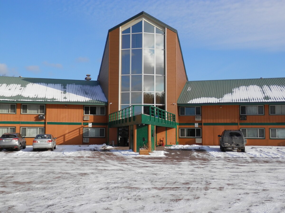 The exterior of a multi-story building is showcased, featuring a slanted roof and a large glass front. Snow covers the ground, and several parked vehicles are visible in front of the entrance, surrounded by a clear, opened space.