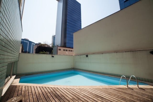 A swimming pool is visible on a wooden deck, offering a clear blue water surface. Surrounding buildings create a modern urban backdrop under a bright sky. The pool area is well-defined, with steps leading into the water and minimal landscaping around.