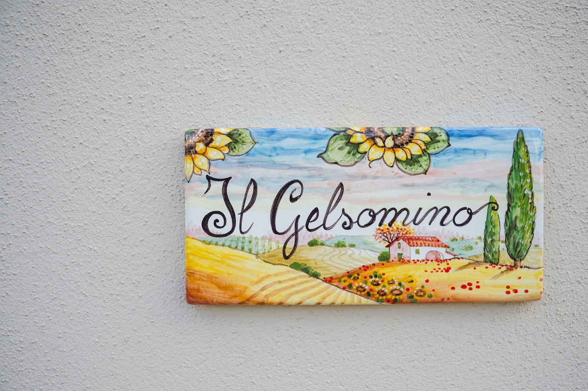 A colorful ceramic sign displays the name 'Il Gelsomino,' featuring hand-painted sunflowers and cypress trees against a backdrop of rolling hills and a distant farmhouse. The sign is affixed to a textured wall, contributing to the rustic aesthetic of the property.