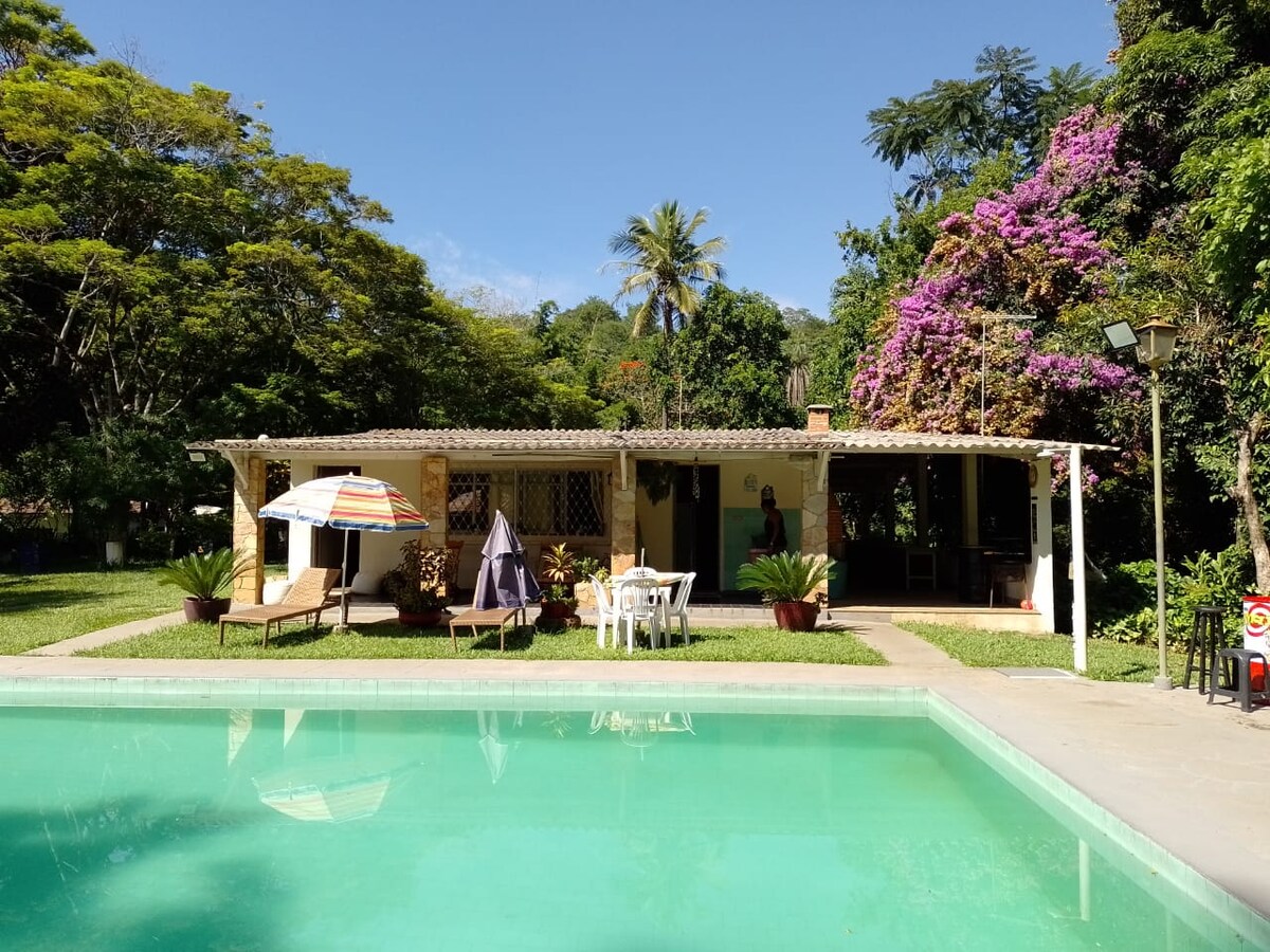 A serene outdoor space features a clear swimming pool with lounge chairs and umbrellas nearby. Lush greenery and vibrant flowering plants surround a charming house, which includes a covered patio area that invites relaxation. Soft sunlight creates a peaceful ambiance.