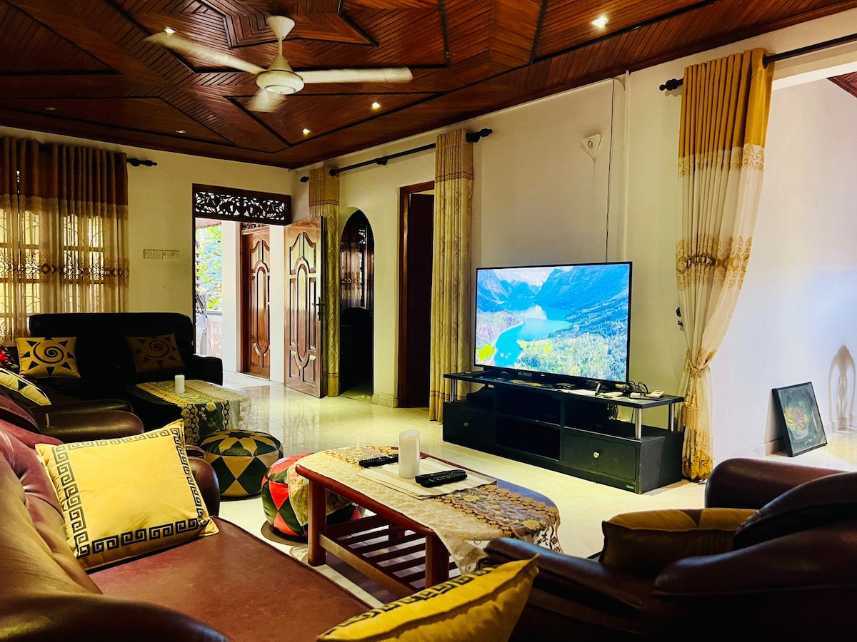 A spacious living area features a flat-screen television positioned on a low black stand. Comfortable sofas are arranged around a coffee table adorned with a decorative cloth. Soft lighting highlights intricate wooden ceiling details and large windows allow natural light to fill the space.