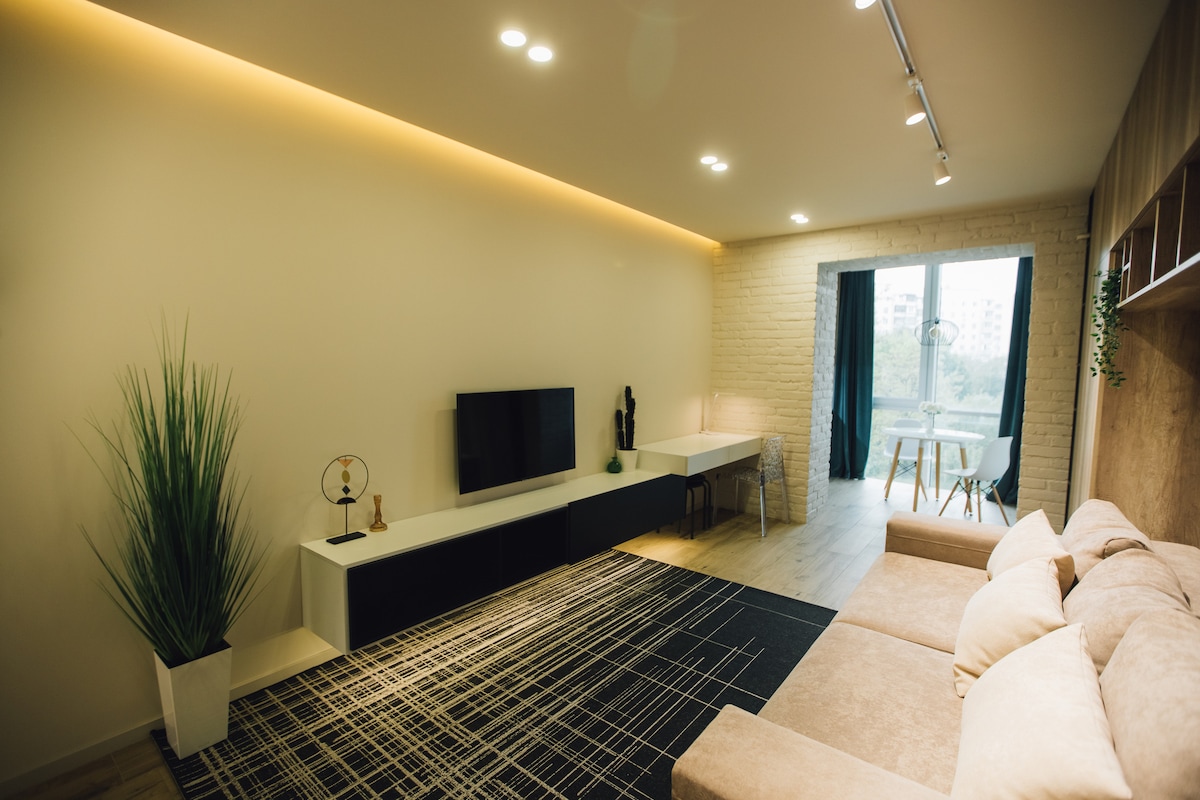 A spacious living area features a comfortable couch and a sleek TV unit against a neutral wall. Natural light floods in through large windows, illuminating a dining table and chairs in the background. Decorative elements, including a floor plant, add a touch of greenery.