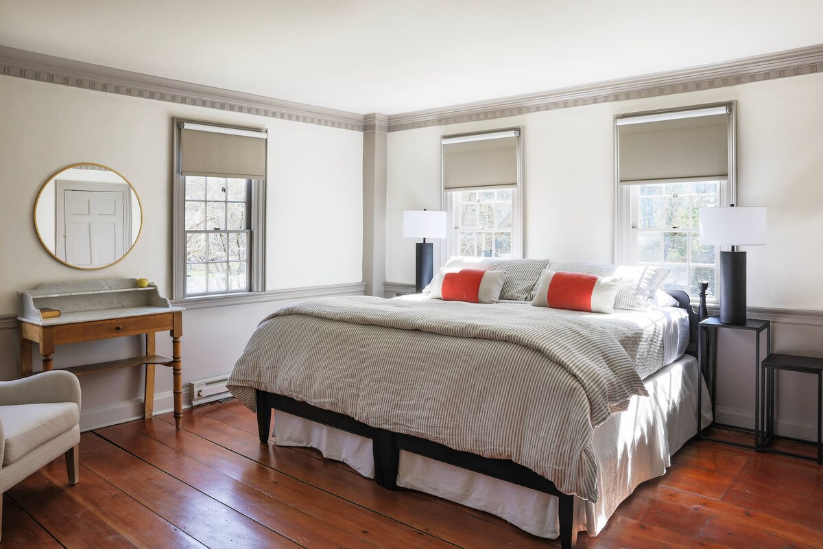 A spacious room features original wood flooring and large windows with soft shades. A king bed with decorative pillows is positioned centrally, complemented by bedside tables with lamps. A round mirror and a small writing desk enhance the room's functionality and comfort.