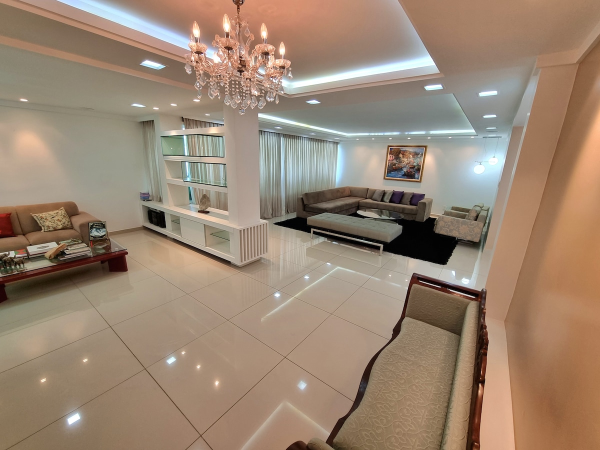 A spacious living area features a modern design with glossy tile floors and chandelier lighting. Plush seating arrangements are positioned around a central coffee table, while shelves display decorative items. Large windows allow natural light to fill the room, enhancing the serene environment.