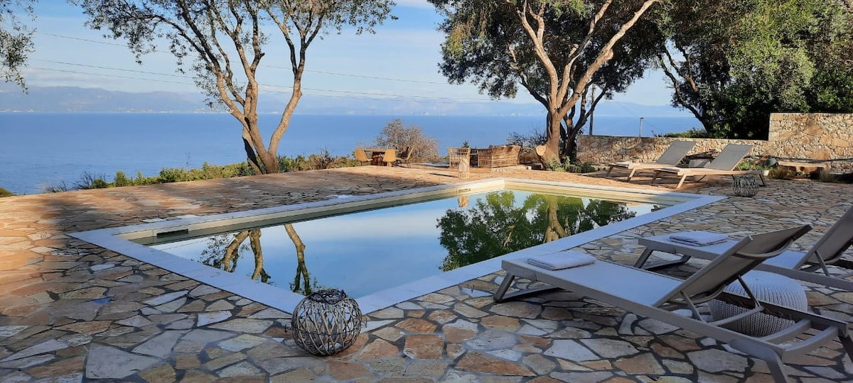 The outdoor area features a serene pool surrounded by natural stone paving, with trees providing shade. Comfortable lounge chairs are positioned nearby, offering a view of the expansive sea and distant hills, inviting relaxation and enjoyment of the scenic landscape.