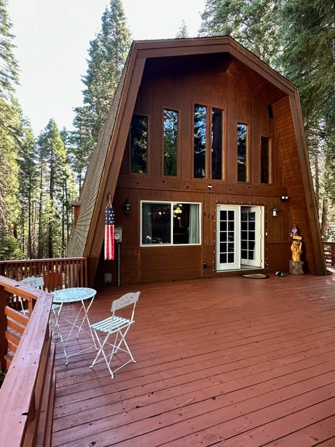 The exterior of the cabin features a wooden façade with tall, narrow windows that allow natural light to fill the space. A spacious deck is visible, adorned with a small table and chairs, surrounded by tall trees that provide a serene backdrop.